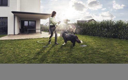 Egy nő a kertjében locsol egy kutya mellett