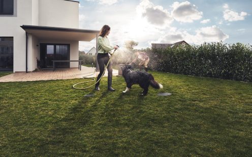 Egy nő a kertjében locsol egy kutya mellett
